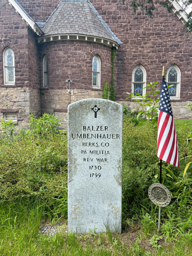 Headstone for a grave marked for Balzer Umbenhauer; Berks CO; PA Militia; Rev War; 1730-1799. In the background, a large church with stained glass windows resides.