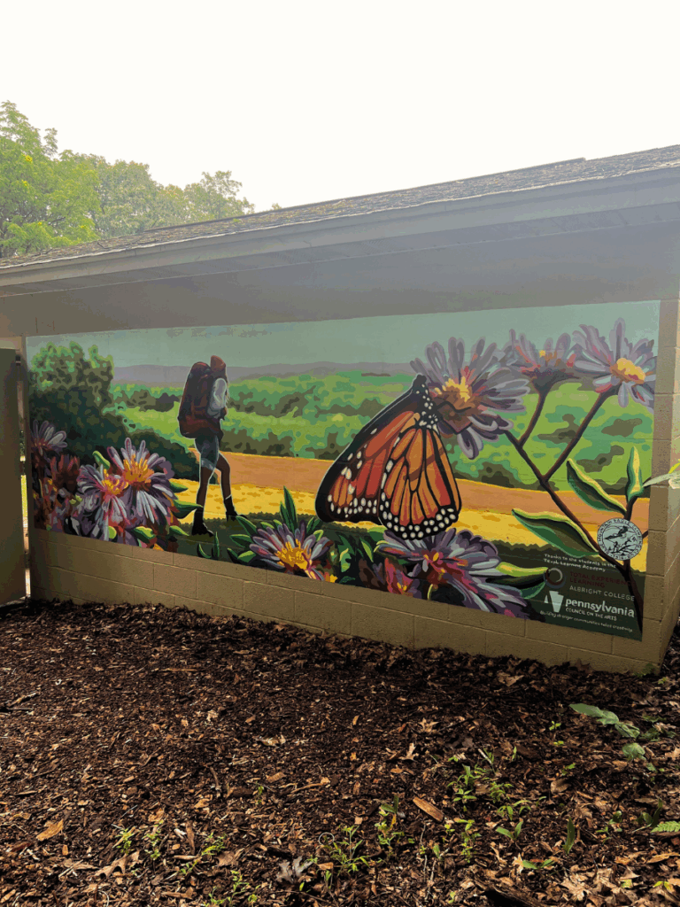 The Wayfinding Mural at Rentschler Arboretum depicts popular flowers and wildlife found on the grounds. Monarch butterflies and a hiker looking off into the mountainous distance are the focus of the mural.
