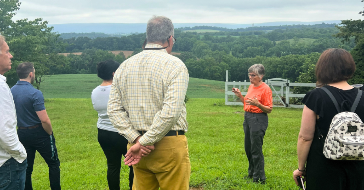 A woman speaks to a group of people with mountains in the background.